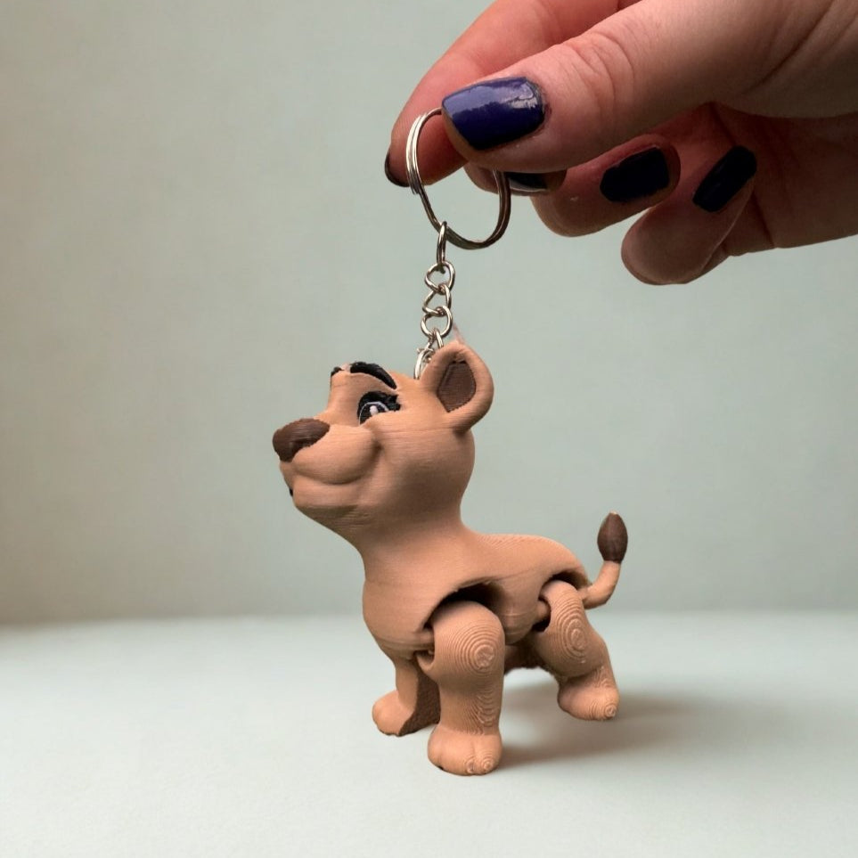 The lion keychains dangle from a set of house keys on a clean white table. The contrast between the plain background and the detailed animal figures draws attention to the craftsmanship and fun character of the design.