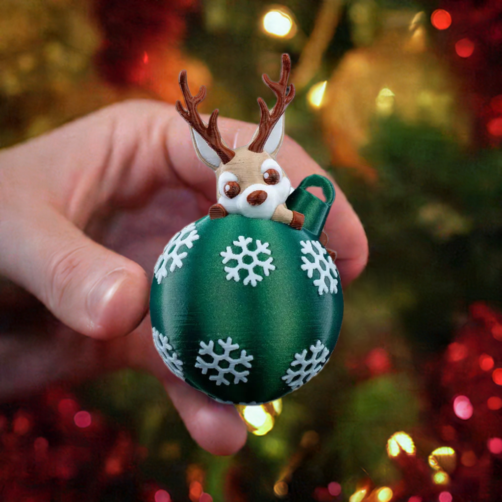 A hand holding a 3D printed ornament featuring a reindeer lying on top of a round green Christmas ornament with white snowflake patterns.