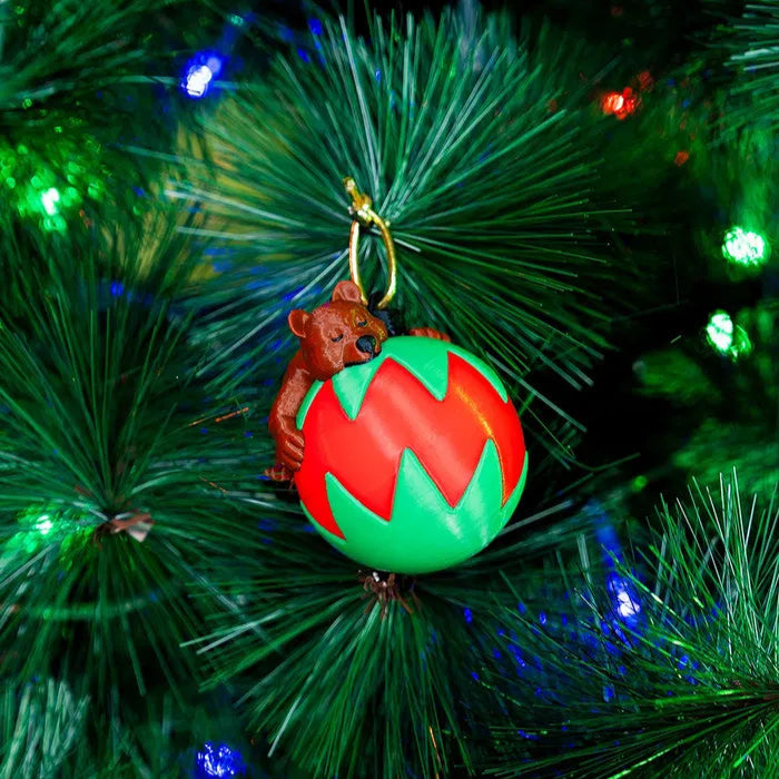 A 3D printed decoration featuring a sleeping bear inside a red and green globe, hanging on a Christmas tree.