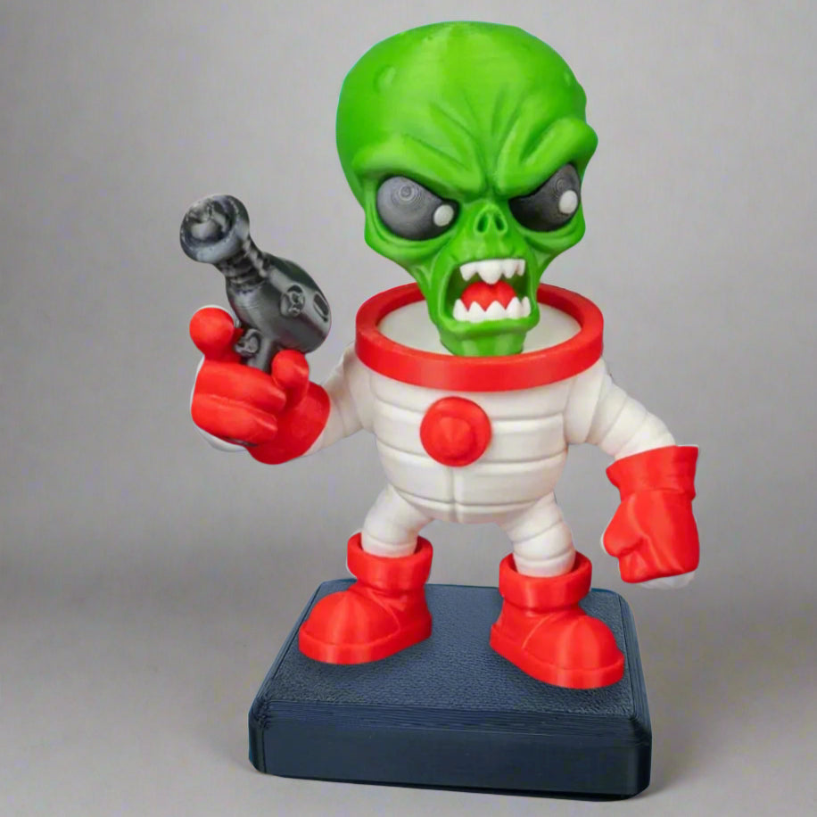 Toy figure of a green alien in a white space suit with red gloves and boots, holding a gun on a gray background.