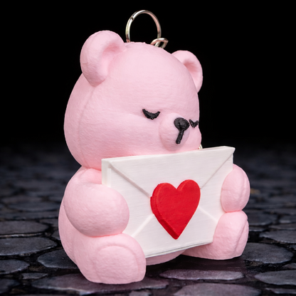 Pink teddy bear keychain holding a heart-shaped envelope on a dark stone surface