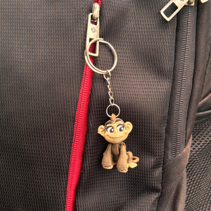 The monkey keychain lies next to everyday objects like coins, keys, and a pen to show scale; its compact size makes it ideal for pockets, bags, or keyrings.