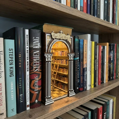 A decorative bookshelf bookend shaped like an arched library aisle with columns and a glowing interior, displayed on a wooden bookshelf between books.
